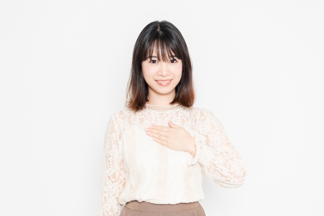 白背景の前に立って、胸に手を当てる、レースのブラウスの女性